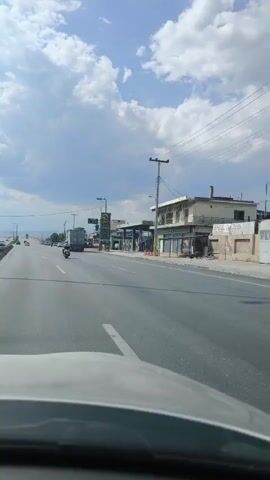 The media captures a dynamic view from inside a vehicle traveling on a highway in Aspropyrgos, Greece. The scene is dominated by a multi-lane road with various vehicles, including cars, trucks, and a motorcycle, navigating the asphalt. The highway is bordered by commercial establishments, notably a gas station with visible price displays showing "1.980" and "1.970". One of the buildings appears to be a business with a signage that includes "ΕΤΕΚΑ GAS". The weather is partly cloudy with a bright sky, suggesting daytime. The overall atmosphere seems to be that of typical road travel on a sunny day in a commercial area. The camera perspective suggests the media was recorded by a passenger or dashcam.
