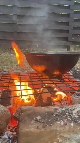 A black metal pot rests on a black metal grate over a small fire pit constructed of red and grey bricks. Flames of red and orange lick the bottom of the pot. Steam rises from the pot as the fire crackles. A grey wooden fence is visible in the background, and green grass is seen on either side of the pit.