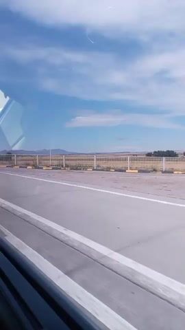 The video shows a roadside scene with a highway or road in the foreground. On the right side of the road, there is a pedestrian overpass with stairs leading up to it. A bus stop shelter is visible next to the overpass. A person is standing at the bus stop. Further back, there are fields and distant hills under a blue sky with scattered clouds. A car is parked on the side of the road. The road has white lane markings and yellow and black curb markings. The video appears to be shot from a moving vehicle.