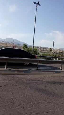 The media file is a video recording from a moving vehicle, likely a car, traveling along a road. The perspective is from the passenger side, looking outwards. The foreground is dominated by the road surface, showing cracks and wear, with a yellow line marking the lane. A metal guardrail with vertical posts separates the road from the area beyond.

The middle ground features a landscape with a variety of trees and vegetation, some appearing manicured, suggesting perhaps agricultural fields or vineyards, consistent with the Charvak, Uzbekistan context. Several utility poles and what appear to be streetlights, some with solar panels, are visible, indicating infrastructure along the roadside. Beyond the vegetation, rolling hills and mountains form the backdrop, their slopes covered in greenery and possibly vineyards.

The sky is a clear, pale blue with a few wispy white clouds, suggesting fair weather and a bright, sunny day. The time of day appears to be late morning or early afternoon due to the clear sky and the quality of light. There are no people visible in the recording. The overall mood is one of travel and observation of the passing scenery, a typical view from a journey through a rural or semi-rural area. There is no visible text in the media file.