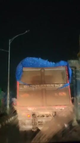 The video shows the rear of a large truck at night. The truck's cargo is covered by a blue tarp. The truck's taillights are illuminated. The ground appears wet, suggesting recent rain or a flooded area. The truck has "BHARATBENZ" written on its back. The view is slightly blurry, as if shot through a wet windshield or from a moving vehicle.