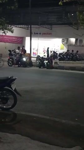 The video shows a street scene at night in front of a store. The store has a sign that reads "luwis pacitan" and appears to be a retail establishment. There are many motorcycles parked in front of the store, suggesting it is a popular place. Several people are visible, some on motorcycles and some walking. One person is riding a bicycle. The street is paved and there is a sidewalk. The lighting suggests it is after dark. There are trees and some overhead structures, possibly awnings or part of the building. The overall atmosphere appears to be one of a typical urban or semi-urban environment in the evening.