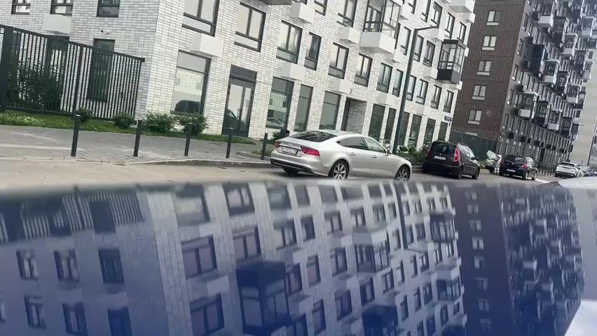The video shows a street scene with modern apartment buildings. In the foreground, the reflection of the buildings is visible on a blue, reflective surface, likely the hood of a car. In the background, several cars are parked along the street. A silver Audi is parked in the middle ground, and other cars are visible further down the street. The buildings have a contemporary architectural style with large windows and balconies. The sky is partly cloudy. The camera appears to be positioned low to the ground, giving a unique perspective of the reflection. No people are visible in the frame.