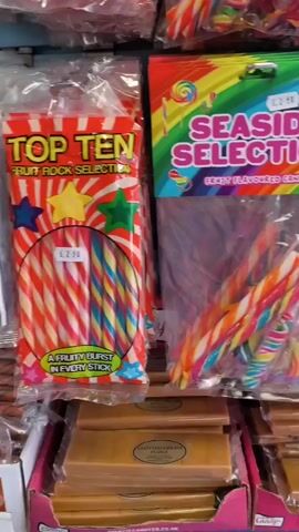 The video shows a close-up view of shelves stocked with various types of candy. In the foreground, there are rows of candy sticks in colorful packaging, some with rainbow designs. To the right, a package labeled "SEASIDE SELECTION" containing several candy sticks and lollipops is visible. Below this, there are rectangular blocks of what appears to be fudge or nougat. Further to the right, a package with the text "TINY TITTIES" and candy shaped like small breasts on sticks is displayed. Next to it, a package labeled "WOBBLY TITTIES" with similar candy is shown. To the far right, a package labeled "WOBBLY WILLIE" containing phallic-shaped candy on sticks is present. The background is blurred but suggests more candy displays. The overall environment appears to be a shop or market stall specializing in novelty candies.