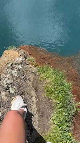 A first-person perspective shot begins looking down at a person's legs and feet, clad in white sneakers, standing on the edge of a rocky, grassy cliff. The cliff overlooks a body of vibrant blue water. The camera then pans up and to the right, revealing more of the blue water, which appears to have some disturbed ripples on its surface, possibly from something dropping in. The scene expands to show a wider landscape: a serene blue pool of water sits in the foreground, with several people gathered on the grassy bank on the opposite side. They appear to be taking pictures of the water and the surrounding scenery. In the background, a lush green valley is cradled by towering, snow-capped mountains under a partly cloudy blue sky. The overall impression is a breathtaking natural landscape with people enjoying its beauty.