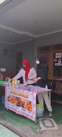 Two women are standing behind a food stall with a pink and white striped banner that reads "Aneka Risoles." The woman on the left, wearing a red hijab and white jacket, is using tongs to place food into a container. The woman on the right, wearing a black hijab and glasses, is also looking at the food. There are several containers of food on the stall, as well as some cans and bottles. The stall is set up outside a building, with a gray wall and a wooden door visible in the background. There is also a fan on the left side of the frame.