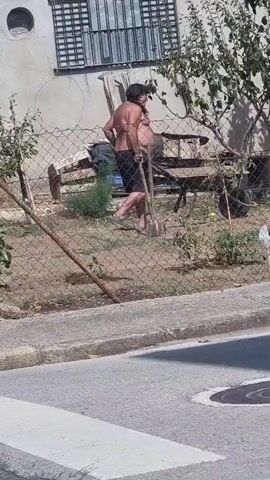 A person is seen in a yard behind a fence, carrying a shovel and a wheelbarrow. The person is shirtless and wearing dark shorts. There is a building in the background and trees and bushes in the yard. The video is filmed from across a street.