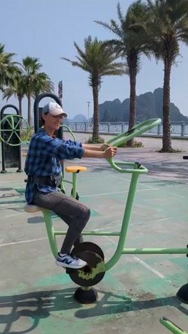 A young adult is using an outdoor exercise bike in a park area. The individual, wearing a blue plaid shirt, gray leggings, and a cap, is seated on the lime-green exercise machine. They are pedaling with their feet and moving the handlebars with their arms, simulating a cycling motion. The exercise equipment is located on a green surface with painted lines, and in the background, there are palm trees and a distant body of water with islands or land masses. The sky is clear and blue, suggesting a sunny day.  The person turns and smiles at the camera briefly.