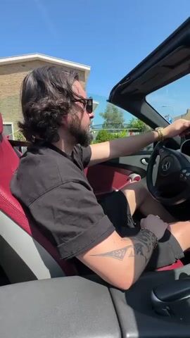 A mid-shot shows a man driving a red convertible. He has dark hair and a beard, and is wearing sunglasses, a black t-shirt, and black shorts. A gold watch is visible on his left wrist, along with a bracelet and a tattoo. The car is a Mercedes-Benz, and the top is down. The surrounding environment consists of houses and trees, with a clear blue sky above. The car is moving at a slow speed, passing by residential buildings.