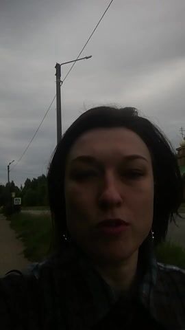 A woman with dark hair and red lipstick smiles at the camera, wearing a brown and blue plaid shirt. She has on dangle earrings. In the background, a sidewalk, a street, trees, a lamppost, and a yellow sign are visible. The sky is overcast. The video is oriented vertically.