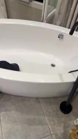 The video shows a bathroom with a white tub that has a black garment inside. The camera pans towards a group of four people standing in front of a mirror. The people are talking, but it is hard to discern what they are saying. One person in a white puffer jacket takes a selfie in the mirror. The bathroom has tan walls, a shower, and a dark stone sink.
