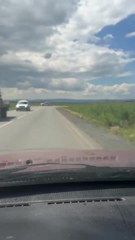 The video shows a view from inside a vehicle traveling on a two-lane road. The sky is partly cloudy with blue patches visible. On the right side of the road, there is a field of tall green grass or reeds. In the distance, there are hills and mountains. Several other vehicles are visible on the road. The dashboard of the vehicle is visible in the foreground. The car appears to be turning to the right.