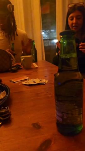 The video shows two people sitting at a wooden table, seemingly playing cards. A man with dreadlocks and a hat is visible on the left side of the frame, and a woman wearing glasses is on the right. Multiple bottles of green beer are on the table, along with playing cards, a mug, a small designer purse, keys, and a black tray with debris. A smartphone is also visible on the table. The background features curtains and a window showing the outside, which appears to be nighttime. Music plays in the background. The man picks up a beer bottle and opens it. Both players seem to be focused on the card game.