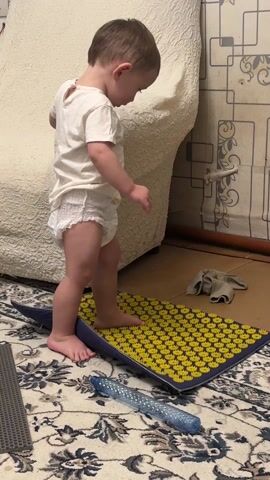 A young child, dressed in a white t-shirt and diaper, is standing on a blue and yellow acupressure mat on a patterned carpet. The child steps on the mat and immediately cries out. The child then steps on the mat again, holding onto the side of an off-white sofa for support. A grey mat and a water bottle are also on the carpet near the acupressure mat. The wallpaper on the wall behind the sofa is geometric and beige.