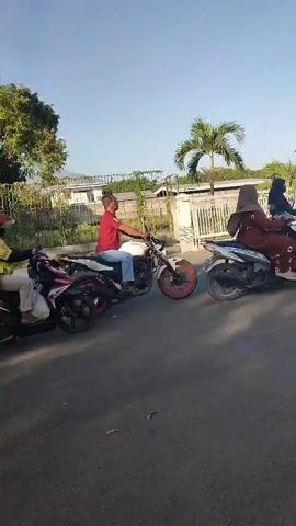 The video shows a line of motorcycles and a few small vehicles waiting at what appears to be a gas station. The motorcyclists are mostly wearing helmets and casual clothing. Some are looking forward, while others are looking at their phones or talking to each other. The environment is a paved road with some greenery and buildings in the background. The sky is clear and sunny. The overall impression is one of a typical day at a busy gas station, with people patiently waiting their turn.