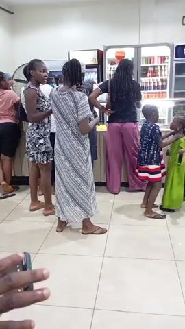 The video shows a group of people standing in what appears to be a food service establishment, possibly a restaurant or cafeteria. There are several people in a line, facing a counter. In the foreground, a person is holding a smartphone, filming the scene. In the middle ground, two young girls are standing near the counter, one wearing a blue and white patterned dress and the other a green dress. Behind them, several adults are interacting with staff behind the counter. The establishment has refrigerators stocked with drinks and displays of food. The floor is tiled, and the lighting is artificial. The overall atmosphere appears to be a busy service environment.