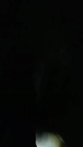 A single headlight illuminates a dark road at night. The shadow of the motorcycle mirror and part of the rider's body are visible in the foreground. The road stretches into the distance, illuminated by the headlight. The wind is blowing, indicated by the noise.