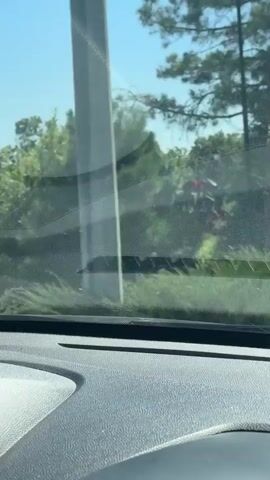 The video shows a view from inside a vehicle, looking out the windshield. The view is slightly distorted by the glass and the glare from the sun. Outside, there are trees, bushes, and some red flowers. The sky is blue and clear. The dashboard of the car is visible in the foreground. The car appears to be moving slowly, possibly in a parking lot or a driveway, as evidenced by the changing scenery and the slight blur of the trees. There are no people visible in the video.