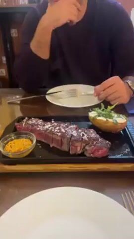 A man wearing a maroon sweater and a watch is sitting at a restaurant table. In front of him is a sizzling cast iron plate with a sliced steak covered in coarse salt, a baked potato with cream and arugula, and a small dish of yellow sauce. The man is using a knife and fork to cut a slice of the steak. There is a small white plate in front of him and a larger white plate at the bottom of the frame. The table is wooden.