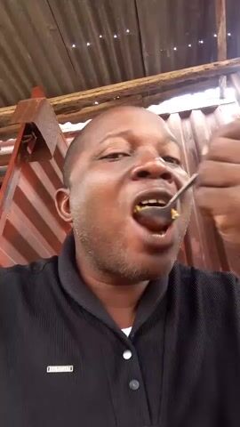 A man with dark skin, a shaved head, and a beard is eating with a spoon from a bowl. He is wearing a black collared shirt. The background consists of corrugated metal walls and wooden beams, suggesting an outdoor or semi-outdoor structure. The lighting is dim and appears to be natural light. The man's facial expression indicates he is actively chewing or tasting the food.