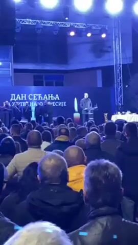 The video shows a nighttime event, likely a commemoration or political gathering, taking place outdoors on a stage. An audience of mostly adult men stands facing the stage. On the stage, multiple people are visible, including a man speaking at a podium, with the words "Dan Secanja" in Serbian script displayed prominently behind him on a large screen. Applause is heard as the camera pans to the large screen, which then shows a close-up of a man's face with some text. A camera operator is seen among the crowd, filming the event. The setting appears to be an urban area at night, with stage lighting and visible building structures in the background.