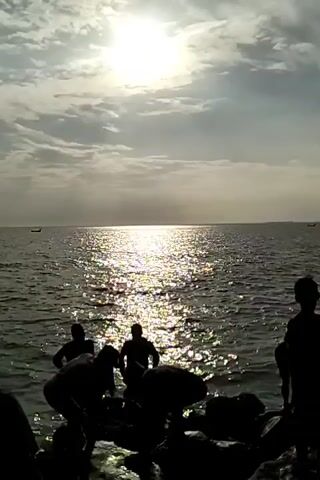 The video shows a group of people silhouetted against a bright, shimmering ocean at sunset. The sun is high in the sky, partially obscured by clouds, and casts a strong glare on the water's surface. The people are standing on a rocky shore, some bending over, possibly examining the rocks or water. In the distance, there are two small boats on the horizon. The overall impression is one of a group of people enjoying the seaside at dusk.