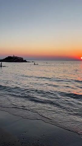 The video shows a beach scene at sunset. The sun is setting over the ocean, casting a warm orange and pink glow across the sky and water. Gentle waves lap at the shore. Several people are visible in the water, swimming or wading. A rocky outcrop with a few people standing on it is visible in the distance to the left. The water is calm and clear, with small waves breaking on the sandy beach.