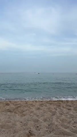 The video opens with a view of a calm ocean under a cloudy sky. The foreground is a sandy beach. Several jet skis are visible in the distance on the water, some with riders and creating wakes. There are also buoys in the water. The scene appears to be a recreational area with water sports taking place.