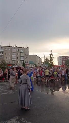 In this vertical, outdoor shot, a crowd of people gathers in what appears to be a town square in Krivodanovka, Russia. The sky above is overcast with a hint of sunset peeking through, suggesting the time of day is late afternoon or early evening. The weather appears to be damp, as the asphalt surface reflects the surrounding buildings and the figures on it.

The scene is dominated by a group of people in the foreground, seemingly engaged in some form of performance or public gathering. Two individuals are dressed in elaborate costumes. One person wears a long, flowing blue robe and a wig that suggests a mythical or fairytale character, possibly a witch or sorceress. Facing this figure is another person in a long, grey dress, also appearing to be part of the performance.

Behind them, a larger crowd of spectators, including many children, watches the proceedings. They are gathered in front of multi-story apartment buildings, typical of Soviet-era architecture, which line the square. A lone, tall evergreen tree stands out against the cityscape. The mood seems to be one of festive anticipation, with the audience appearing engaged and curious. A sign is partially visible on one of the buildings, reading "MARKET." The overall atmosphere is that of a community event, possibly a local festival or a children's performance.