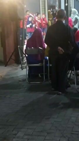 A group of children in red shirts and headscarves stand on a stage or raised platform. A woman wearing a purple headscarf and blue pants sits in a chair facing away from the camera. To her right, a man in a black shirt and pants stands with his hands behind his back. The ground is paved with interlocking bricks. The scene appears to be outdoors at night, with artificial lighting visible.
