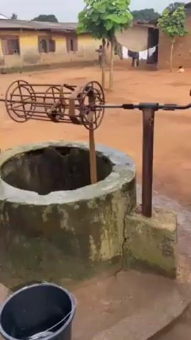 A water well stands in a courtyard in what appears to be a village setting. The well is constructed of concrete and has a dark opening with water visible inside. A rusty, metal hand-crank mechanism for drawing water is positioned above the well. A young person in a yellow shirt begins to crank the wheel. Buildings with tin roofs and windows can be seen in the background, some painted pale yellow. There are also trees with large green leaves visible. A black bucket sits on the ground near the well. The ground is red clay.  Clothes are hanging on a clothesline in the background. The sky is overcast.