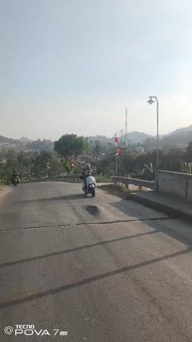 The video begins with a wide shot of a paved road with a slight incline. On the road, a white scooter with a rider is moving away from the camera. The rider is wearing a helmet and appears to be going downhill. To the left of the road, there are some trees and vegetation, and beyond that, houses and buildings can be seen. There are also some red and white flags visible on the left side of the road. To the right of the road, there is a concrete barrier. In the background, there are hills and a clear sky. As the video progresses, the scooter continues to move down the road and out of frame. Another scooter is visible further down the road on the left side. The camera is mounted at a low angle, suggesting it may be attached to another vehicle or held by someone close to the ground. The lighting suggests it is daytime, with the sun likely to the right and slightly behind the camera.