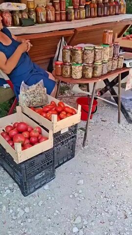 The video shows an outdoor market stall. On the left, a person wearing a blue dress is seated, partially visible. In front of the stall are two wooden crates filled with red tomatoes, and to the left of them is a bag of dried herbs. The stall itself is set up with a wooden counter on the upper level displaying rows of glass jars filled with various preserved goods, possibly pickles and jams. A white lace cloth is draped over the upper counter. Below this, a folding table is covered with a floral tablecloth. This table holds more jars of preserved items, stacked in several rows. A red bucket is visible beneath the table. To the right of the table is a digital scale. Another wooden crate, this one containing peaches, is positioned on the table further to the right. The background consists of trees and greenery, suggesting a rural or semi-rural setting. The ground is covered in gravel. A white pole, likely supporting the market canopy, runs vertically through the center of the frame. The lighting suggests it is daytime.