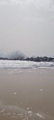 The video shows a beach with dark rocks in the water. Waves are crashing against the rocks and creating a lot of white foam. The sand is light brown and looks wet. The sky is overcast and grey. There is no one visible in the video.
