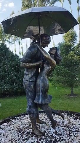 This is a close-up, vertical shot of a bronze statue depicting a couple embracing under an umbrella. The statue is positioned outdoors, surrounded by greenery and a gravel path. The statue appears to be made of bronze and has a weathered, aged patina. The couple is in a romantic pose, with the man holding the umbrella over both of them and the woman leaning into him. The umbrella is tilted, and it appears to be raining, as evidenced by water droplets visible on the umbrella and in the air around the statue. The background consists of trees and a cloudy sky.