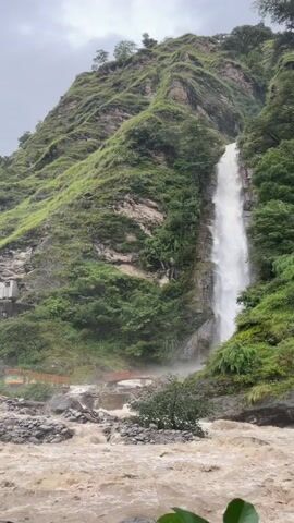 A powerful waterfall cascades down a steep, lush green mountainside. The water is white and foamy as it plunges into a turbid, fast-flowing river below. The riverbed is rocky, with large boulders visible. On the left side of the scene, a small structure or building is built into the mountainside, and a bridge or walkway is visible crossing the river. The sky is overcast, suggesting a cloudy or rainy day. The overall impression is one of raw, natural power and a somewhat wild, untamed environment.