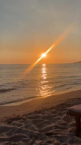 The video shows a serene beach scene at sunset. The sun, a bright orb low in the sky, casts a golden glow across the ocean. Sunlight reflects off the gentle waves, creating a shimmering path on the water. The beach in the foreground is covered in sand, with the edge of a lounge chair visible on the right side. A few distant figures can be seen in the water. The sky is a warm gradient of orange and yellow, with hints of blue at the top. The overall atmosphere is peaceful and tranquil.