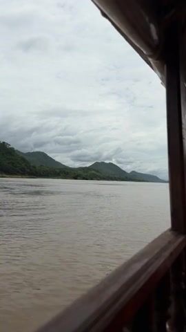 The video shows a view from a boat on a wide river. The river is brown and choppy. In the distance, there are green, tree-covered hills under a cloudy sky. The foreground is dominated by the wooden railing of the boat, which is dark brown. The camera is stationary for the duration of the video.