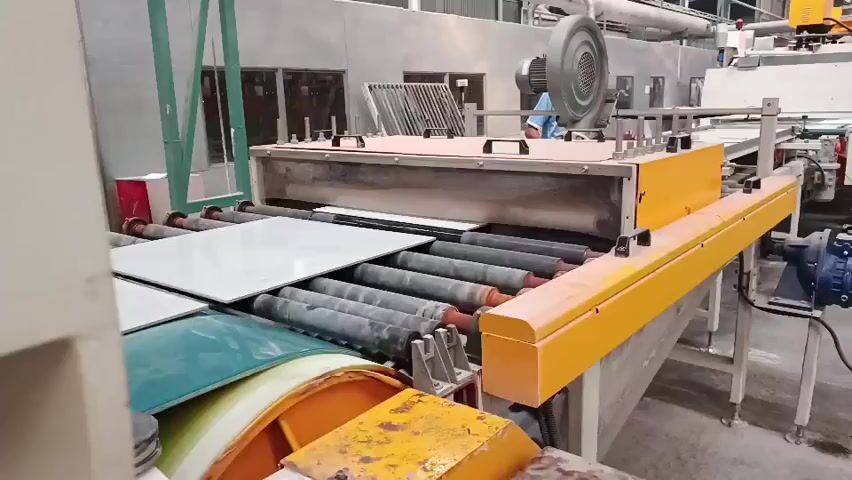 A conveyor belt system moves large white tiles through an industrial setting. The tiles are being processed by a machine with a yellow housing and a silver upper section, which appears to be a type of oven or dryer. Several rollers are visible beneath the tiles, supporting their movement. In the background, two individuals in blue shirts are visible, suggesting human operation or supervision of the machinery. The environment seems to be a factory floor, with pipes and industrial equipment in the background.