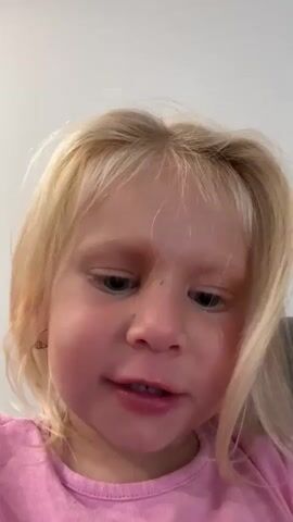 This is a close-up, low-angle video of a young Caucasian girl with blonde hair and blue eyes. She is wearing a bright pink t-shirt. Her mouth is open and she appears to be speaking or singing. She has a small, light-colored earring in her left ear. The background is out of focus and appears to be a neutral-colored wall, suggesting an indoor setting. The lighting is bright and even, making it difficult to determine the time of day. There is no visible text in the video. The girl's expression is animated and engaged, suggesting she is enjoying herself. The setting and her attire do not provide specific clues to a particular location within Milton Keynes, but the indoor setting is evident.