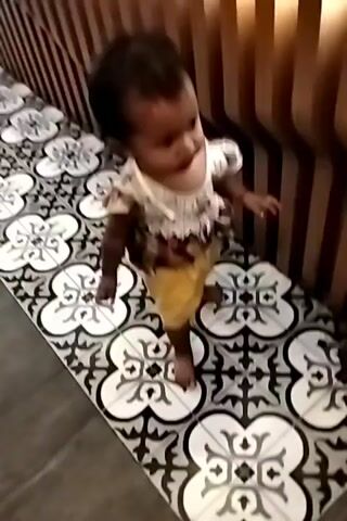 A young child with dark hair, wearing a short-sleeved shirt and yellow shorts, walks across a patterned tile floor. The child turns and walks towards a seating area with wooden chairs and tables. Another child, partially visible, is also present in the background. The environment appears to be an indoor space, possibly a restaurant or cafe. The video is shaky and the image quality is low.