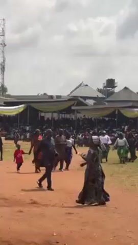 The video shows a large crowd gathered on a grassy field, possibly for an event or ceremony. There are structures covered with yellow and white draped fabric behind the crowd, suggesting a stage or seating area. People are walking across the field, some dressed in traditional attire. A red carpet is visible in the center. The overall atmosphere seems lively, and the event appears to be well-attended. The weather is partly cloudy, and the scene is set outdoors in a location with trees and buildings in the background.