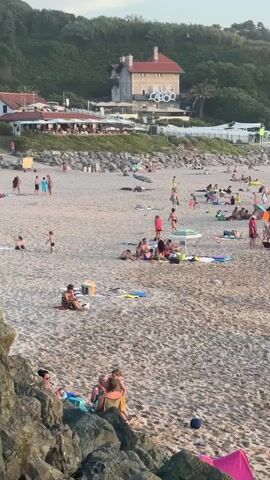 The video shows a crowded beach with many people relaxing, sunbathing, and interacting. The sand is coarse, with scattered rocks and debris. Several beach umbrellas and towels are visible, along with surfboards and cooler boxes. People are dressed in swimwear, and some are walking or playing on the beach. In the background, a building with "BLUE LAGOON" written on it is visible on a hillside covered with vegetation. The sky is clear, and the lighting suggests it is late afternoon or early evening. The overall scene depicts a lively and busy beach environment.