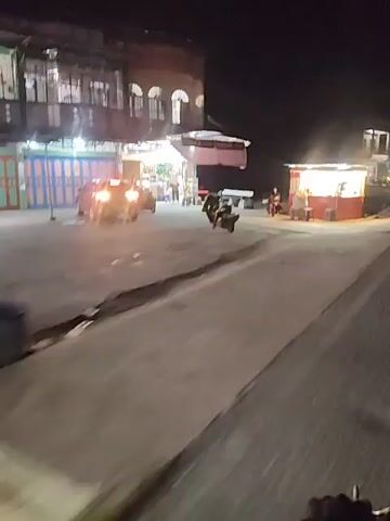 The video shows a nighttime scene in what appears to be a roadside area. The footage is shaky, suggesting it was recorded from a moving vehicle. On the left side of the frame, there's a building with arched windows and bright lights illuminating the storefronts, possibly shops or restaurants. Several motorcycles are parked or moving in front of this building. To the right, there's a small, brightly lit food stall with red trim, and a few people are visible around it. The road in the foreground is paved, and beyond that, there's a ditch or a channel with some debris or vegetation. Further in the background, more buildings and lights are visible, indicating a populated area. The overall impression is a lively street scene at night, likely in an urban or semi-urban environment.