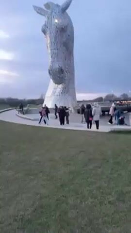 The video shows two large metal horse head sculptures, with a number of people walking around them. The sculptures are situated in a park-like environment with grassy areas, water features, and walkways. The sky is overcast. The camera pans from left to right, showcasing both sculptures and some of the surrounding area. The people appear to be tourists or visitors, observing and interacting with the sculptures.