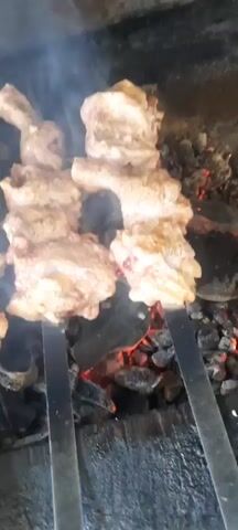 The video shows skewers of meat cooking over hot coals. Smoke rises from the meat and coals, indicating the cooking process. The meat appears to be cut into bite-sized pieces and is seasoned. The background is dark, suggesting the video was filmed at night or in a dimly lit area. The camera angle is close-up, focusing on the food as it cooks.