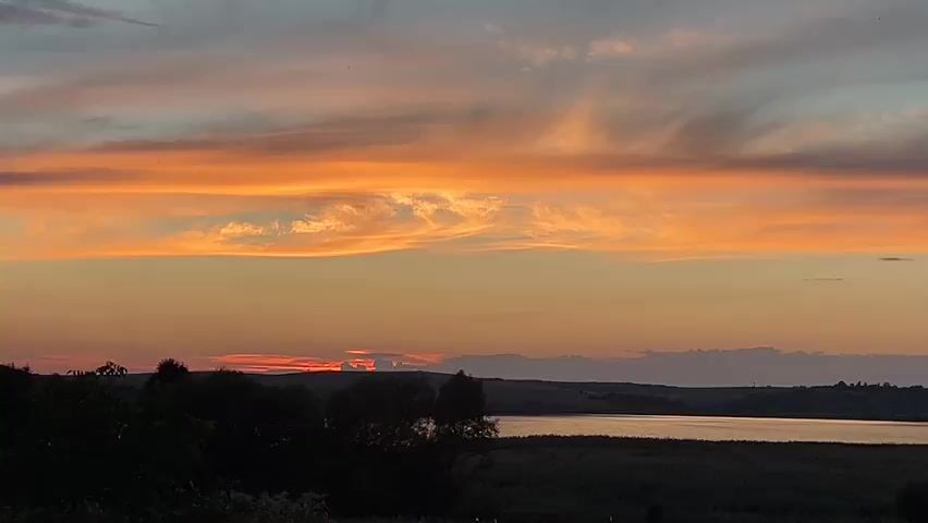 The video shows a sunset over a landscape. The sky is filled with vibrant orange and yellow clouds. Below the sky, there is a dark silhouette of trees and hills. In the distance, a body of water is visible, reflecting the colors of the sunset. The overall scene is peaceful and serene.