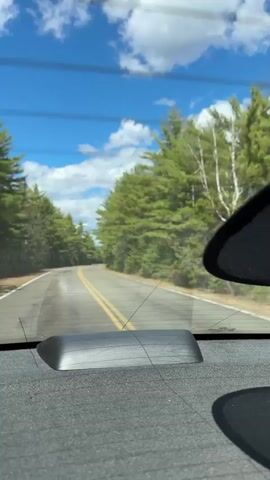 The video shows a view from inside a car driving down a paved road. The road has a double yellow line in the center and is flanked by green trees on both sides. The sky is blue with scattered white clouds. A portion of a dark object is visible in the right side of the frame. The interior of the car is visible, with a gray dashboard and a small black rectangular object near the base of the windshield. The car is moving forward.