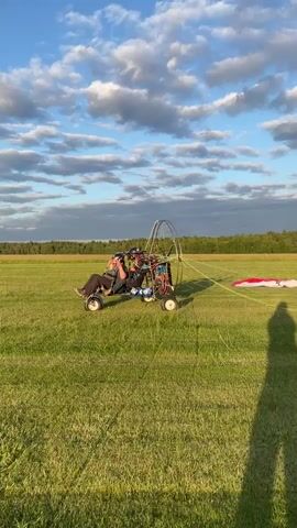 A powered parachute sits on a grassy field under a blue sky with white clouds. Two people are seated in the parachute. The person in front is steering, and the person behind appears to be a passenger. The parachute has a large wing behind it, and a propeller is visible. In the background, a line of trees forms the horizon. A shadow of a person is cast on the grass to the right of the frame. The video is shot from a low angle, looking slightly upwards at the parachute.