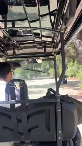 The video begins with a view from inside a golf cart. A person wearing glasses and a white shirt is driving the cart. The cart is moving along a dirt path. Lush green trees and bushes are visible on either side of the path. In the background, a building with a white roof can be seen through the trees. The camera then pans to show more of the surrounding environment, which appears to be a wooded area with a clear blue sky. The golf cart continues to move forward, and the scene transitions to a view of the path ahead, framed by tall trees.
